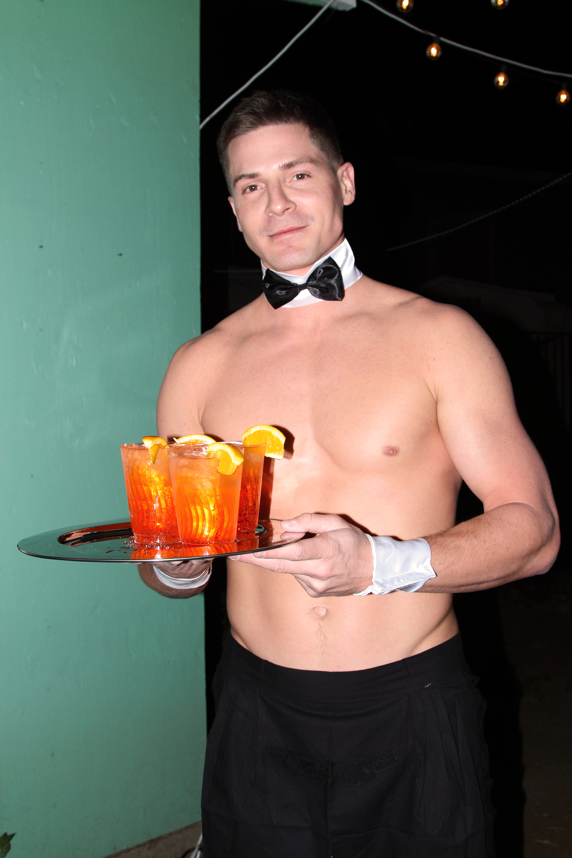 Shirtless white male butler facing the camera with a tray of orange cocktails